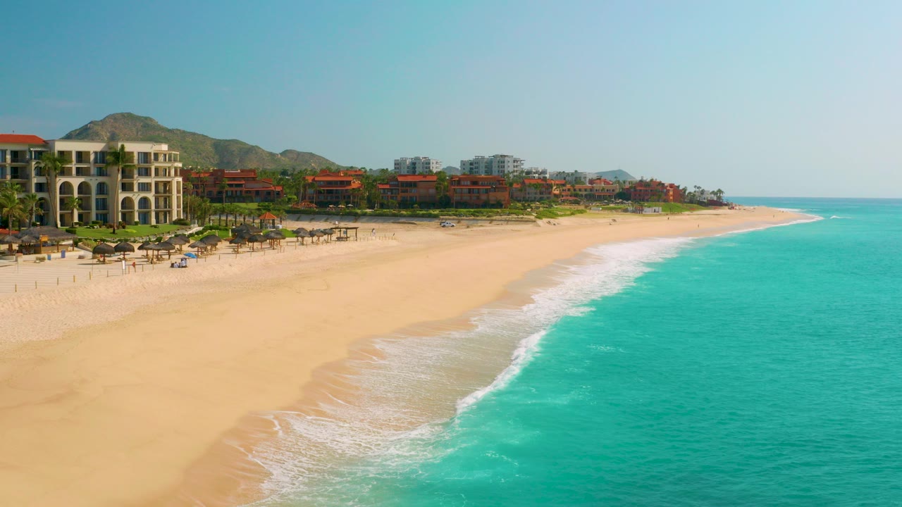 Opening aerial frame over Dreams Beach in Baja California Sur, Mexico, showing pale sand, turquoise ocean, resort buildings, palms, hills, and no visible people.