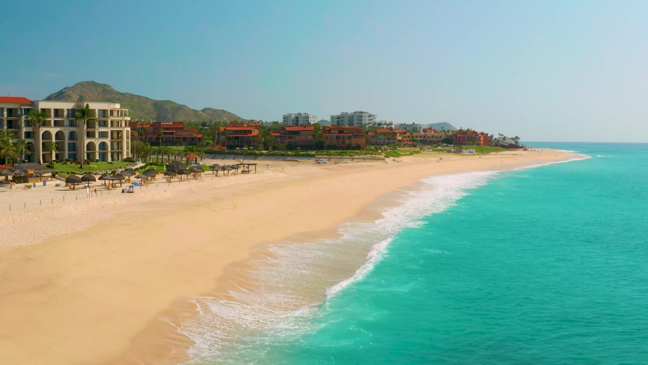 Aerial frame at 0:01 over Dreams Beach, Mexico, with the surf line slightly farther along the sand, resort blocks at left, hills behind, and no visible people.