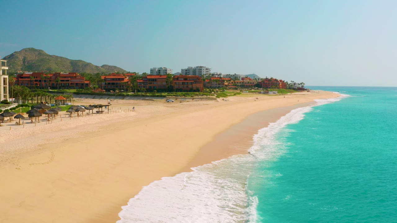 Wide aerial frame of Dreams Beach in Baja California Sur, Mexico, with red-roof resort buildings below green hills, open sand, turquoise surf at right, and small visible beachgoers near shade structures.