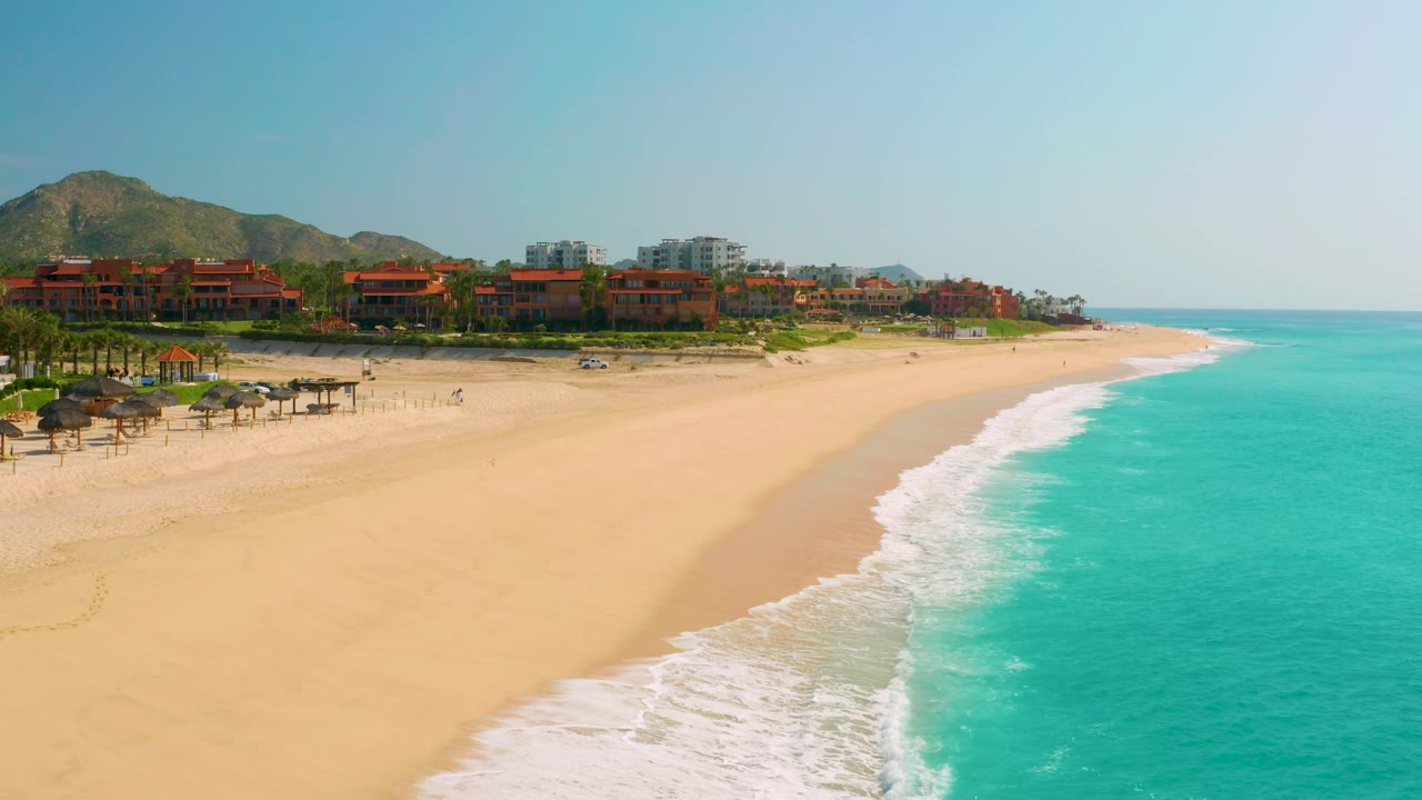 Dreams Beach in Baja California Sur, Mexico appears from above with a broad empty stretch of sand, white foam along the right side, resort buildings and hills behind, and small beachgoers visible near the left beach amenities.