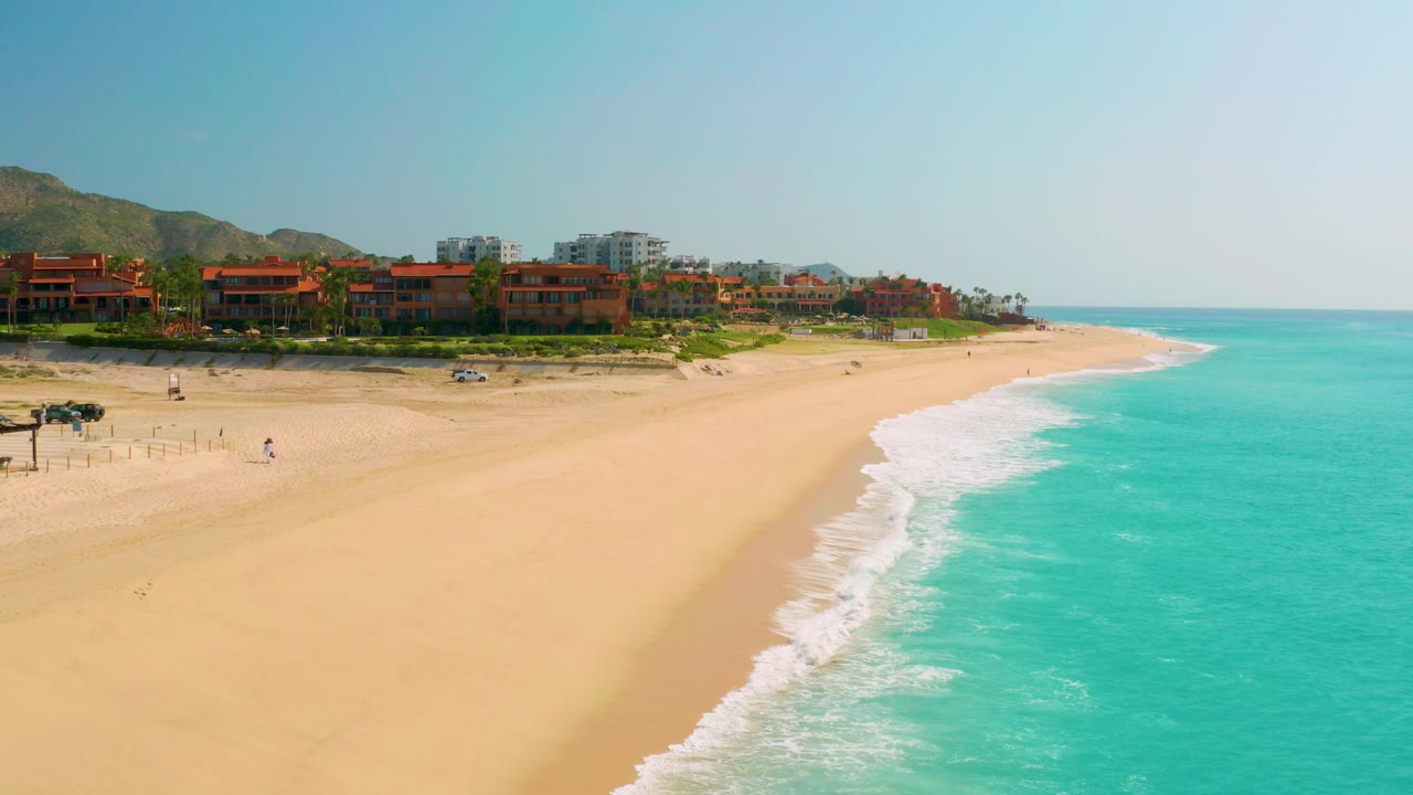 Opening frame of Dreams Beach in Baja California Sur, Mexico, showing a wide sandy shoreline, turquoise water on the right, white surf, beachfront buildings below hills, a parked car near the sand, and no visible people.