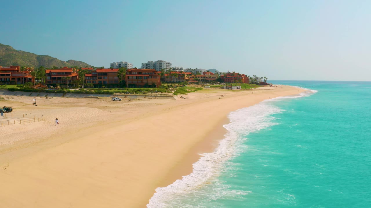 A slightly later view of Dreams Beach in Baja California Sur, Mexico, showing more water to the right, a long white surf edge, tan sand, beachfront buildings at the top of the beach, hills behind them, and no visible people.