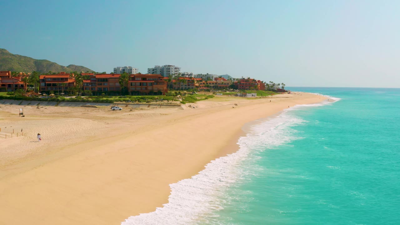 Wide beach and surf with beachfront buildings, hills, clear sky, and a parked car near the sand.