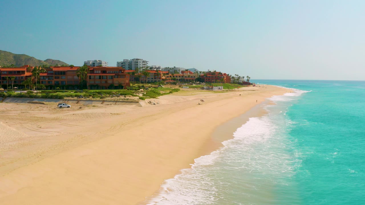 Wide aerial frame of Dreams Beach in Baja California Sur with turquoise water on the right, pale sand in the center, resort-style buildings at left, distant mountains, and no visible people.