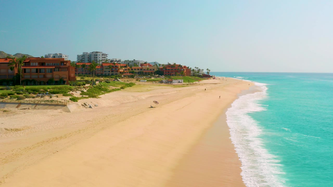 Wide aerial view of Dreams Beach in Baja California Sur with resort buildings along the left edge, pale sand through the center, and turquoise water on the right.