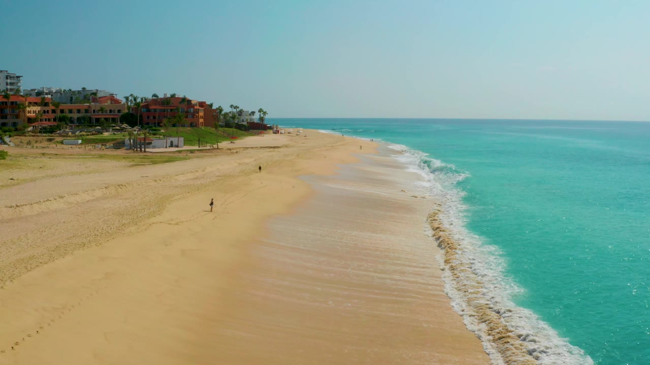 Wide aerial view of Dreams Beach in Baja California Sur, Mexico, with the surf line shifted farther across the frame, resort-style buildings at upper left, and tiny distant people visible on the open sand.