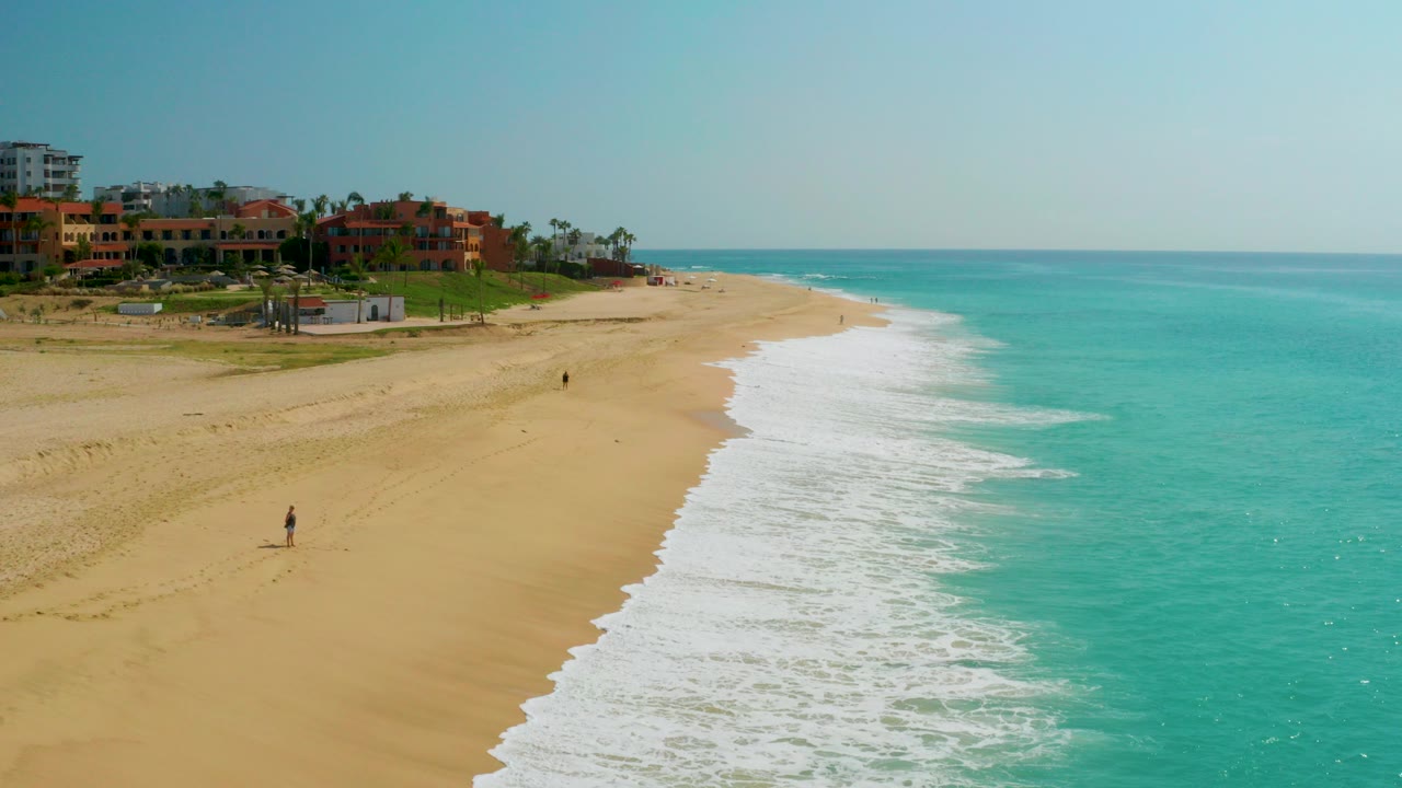 Opening frame of Dreams Beach in Baja California Sur, Mexico, showing a broad sandy shoreline, turquoise water, white surf, resort buildings, palms, and tiny distant beachgoers.