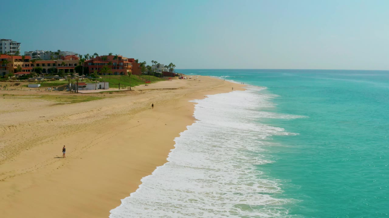 A wide frame of Dreams Beach shows the surf washing up the sand, turquoise sea across the right half, low resort buildings and palms at left, and a small distant walker near the beach.