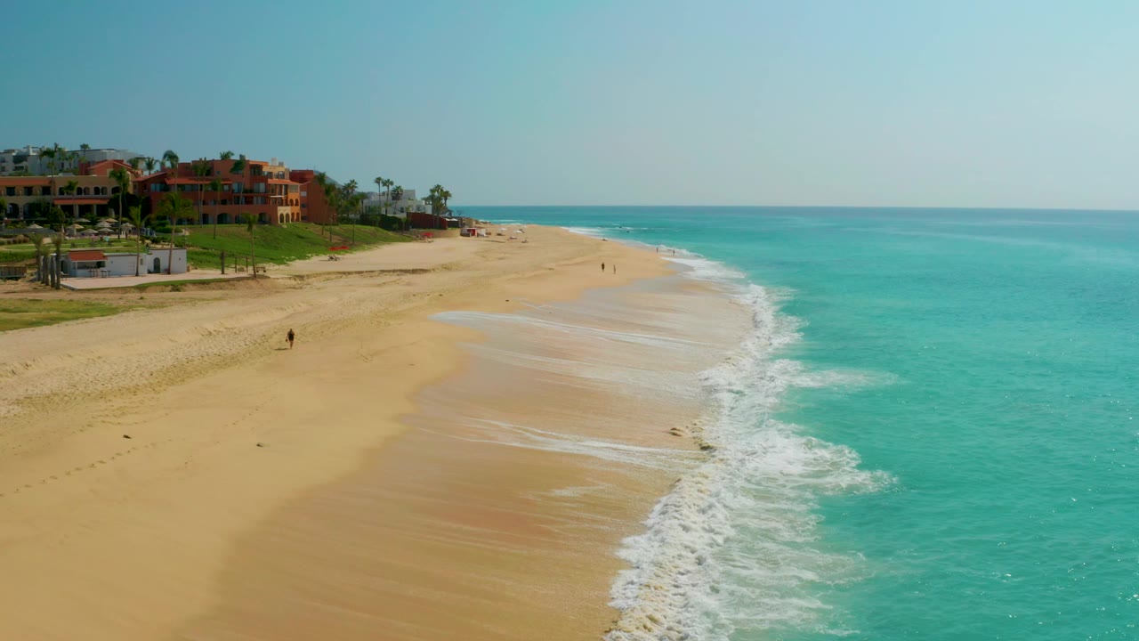 Dreams Beach shoreline from above, with more ocean filling the right side, a bright white surf band along the sand, and red-orange buildings set back at the upper left.