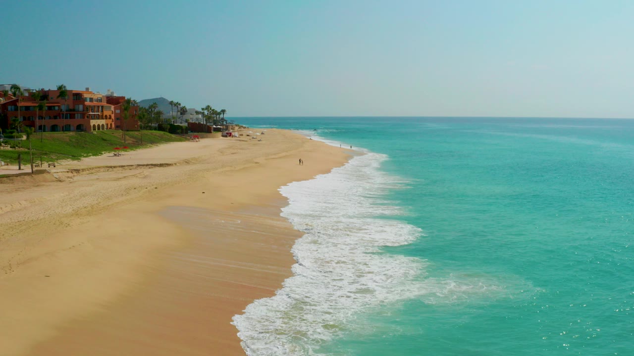 Opening frame over Dreams Beach in Baja California Sur, with turquoise water on the right, pale sand on the left, low shoreline buildings in the distance, and tiny people visible on the beach.