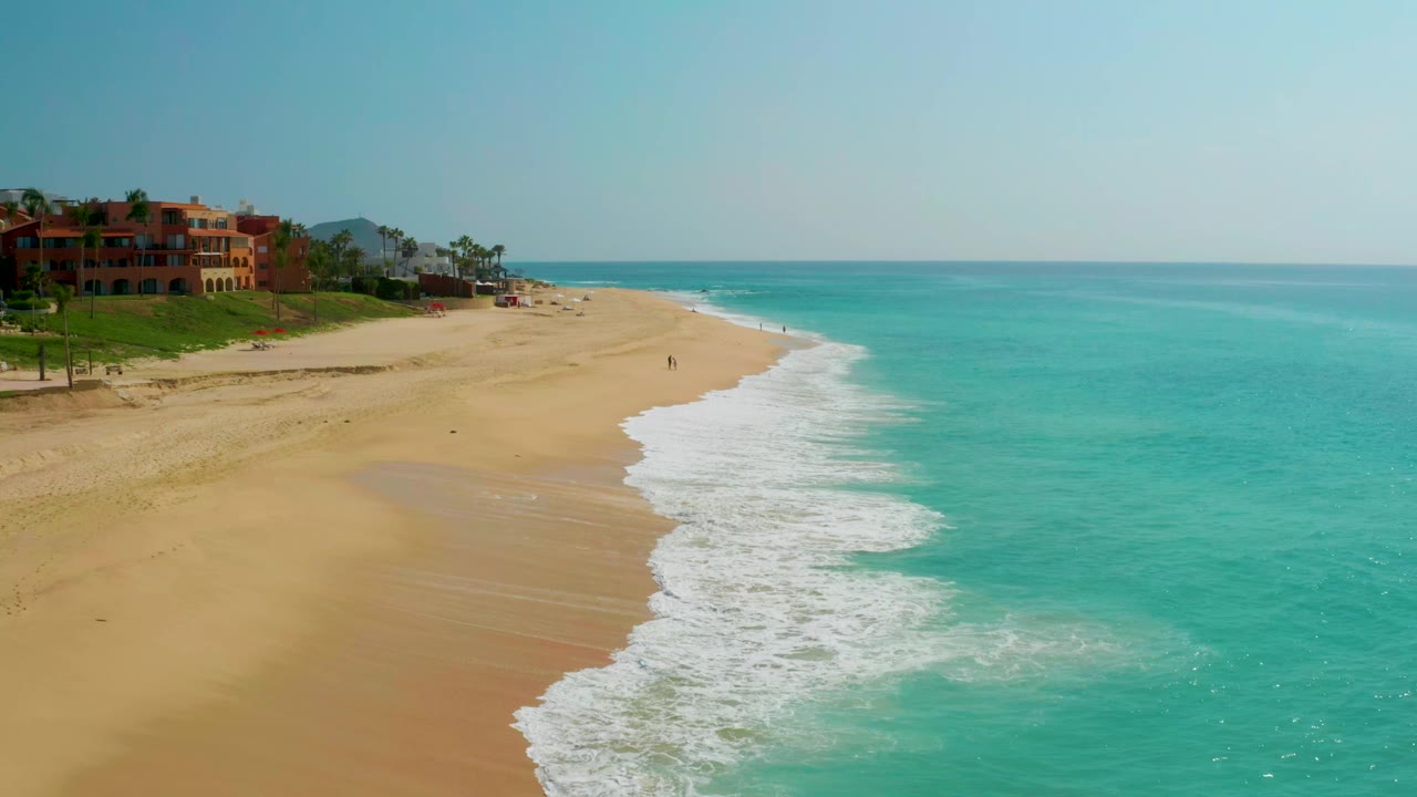 Blue water foams onto a wide sand beach beside distant coastal buildings.