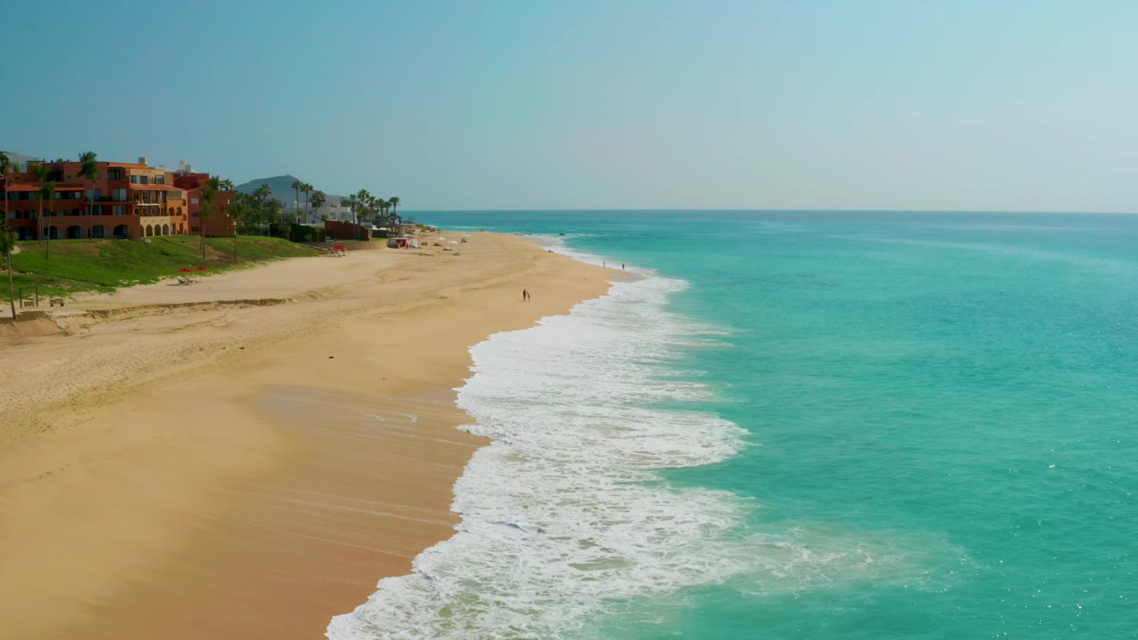 Aerial view at Dreams Beach with the white surf line curving from lower center toward distant sand, turquoise ocean to the right, buildings at far left, and tiny beachgoers visible.