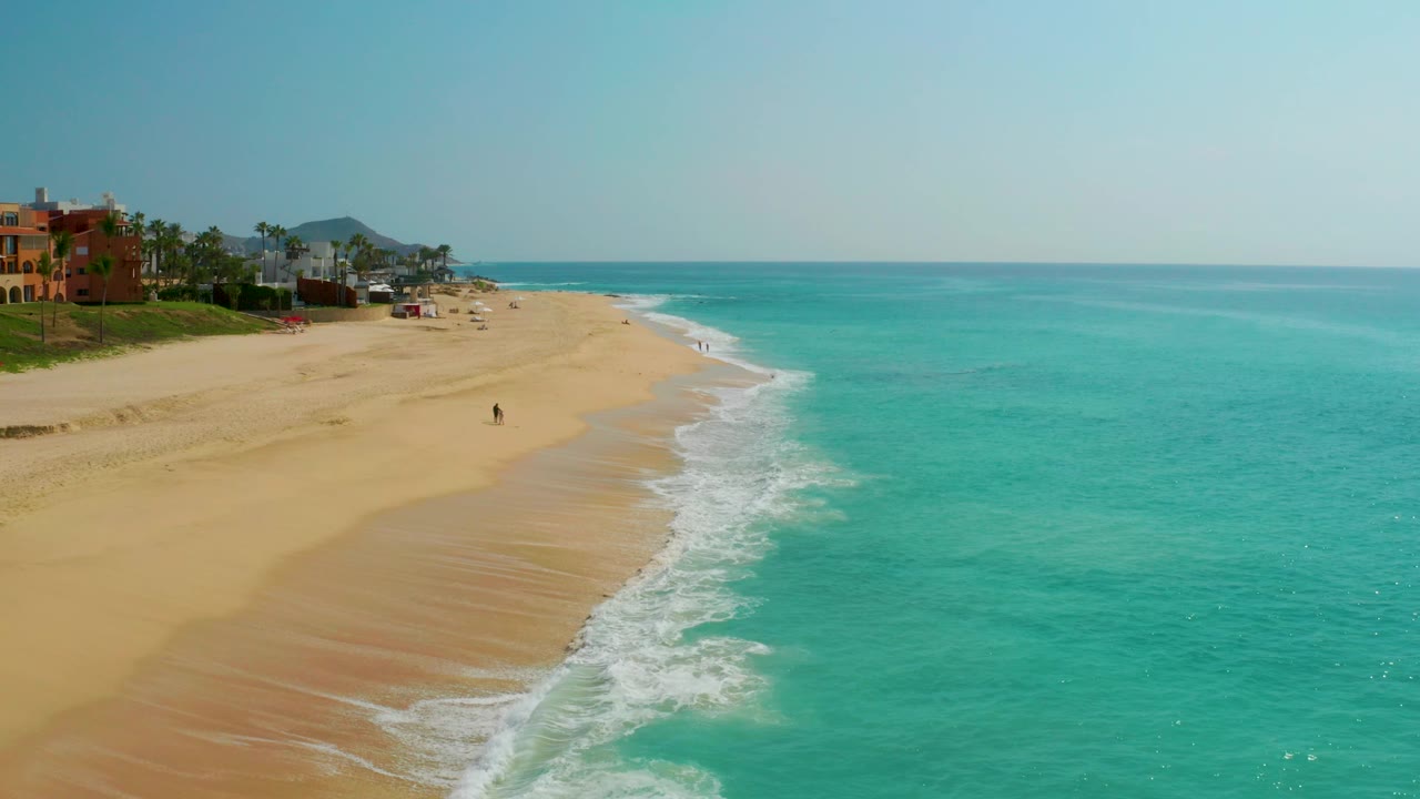 Elevated view over Dreams Beach, Baja California Sur, with turquoise sea filling the right side, pale sand on the left, surf curling at the edge, and no clearly visible people.