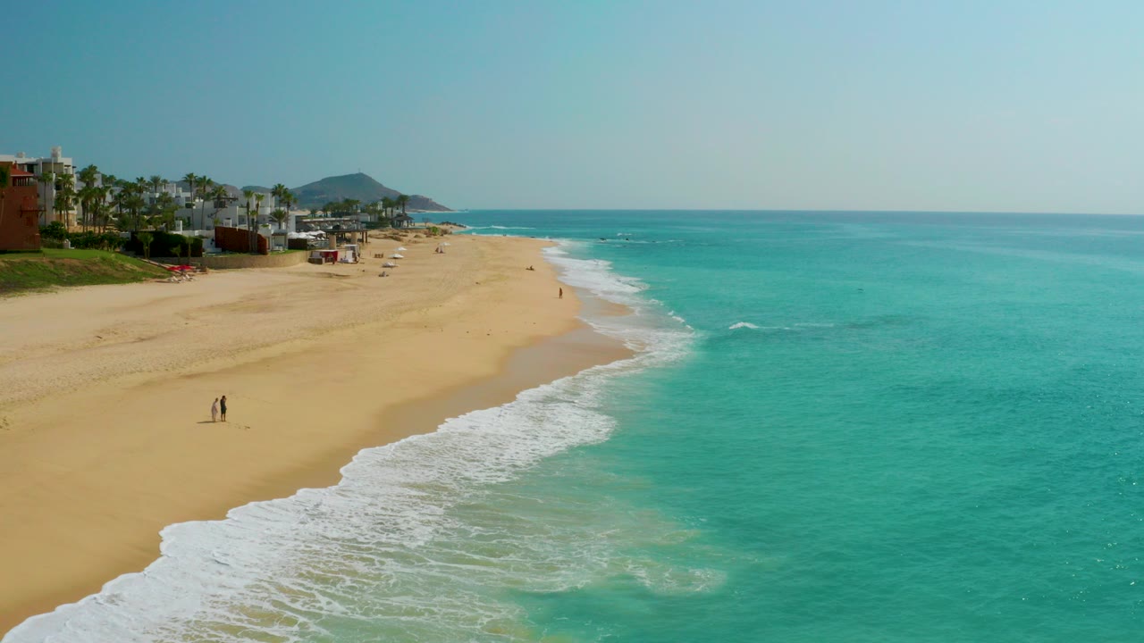 Wide aerial view at Dreams Beach in Baja California Sur, Mexico, with turquoise water on the right, a curving sandy beach on the left, resort buildings in the distance, and tiny people visible on the sand.