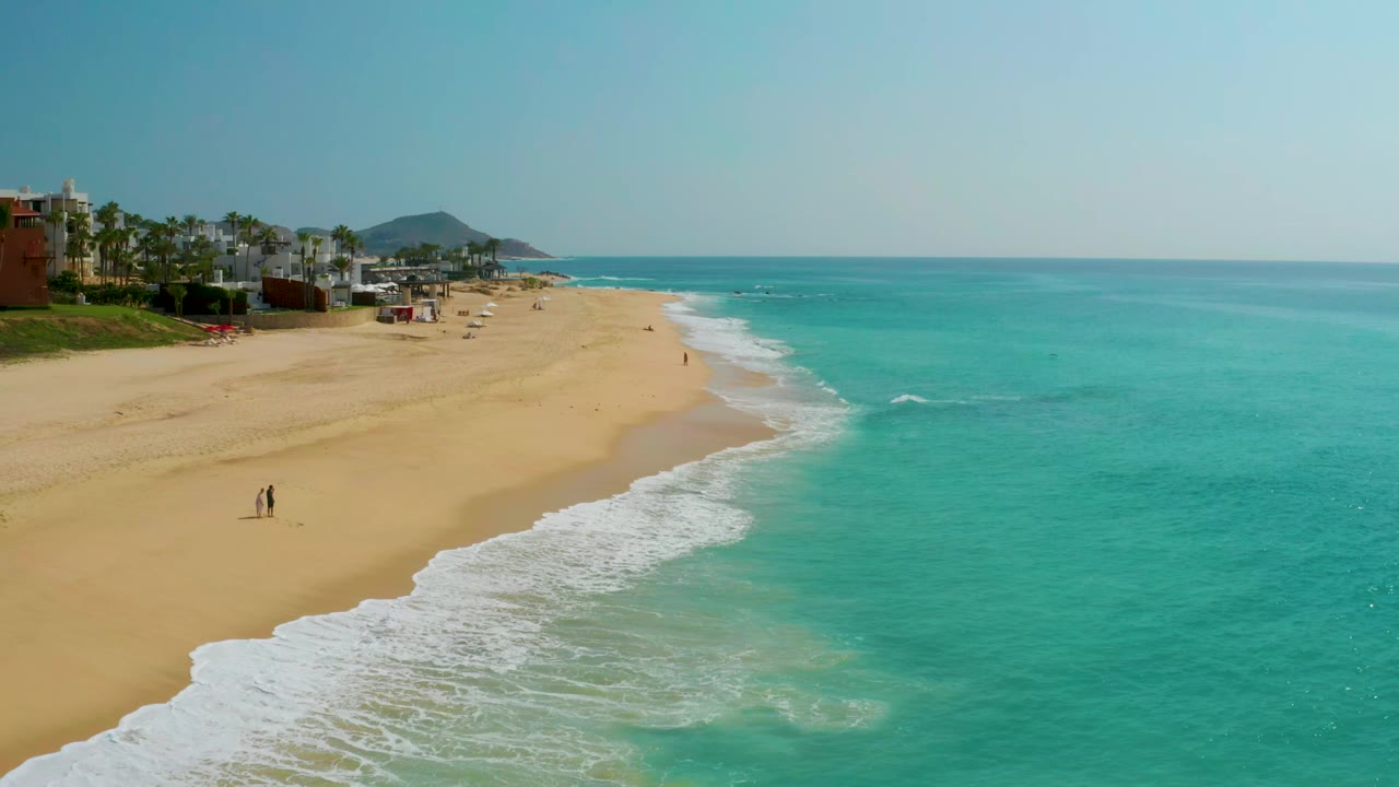 Curving beach with white waves and resort buildings in the background.