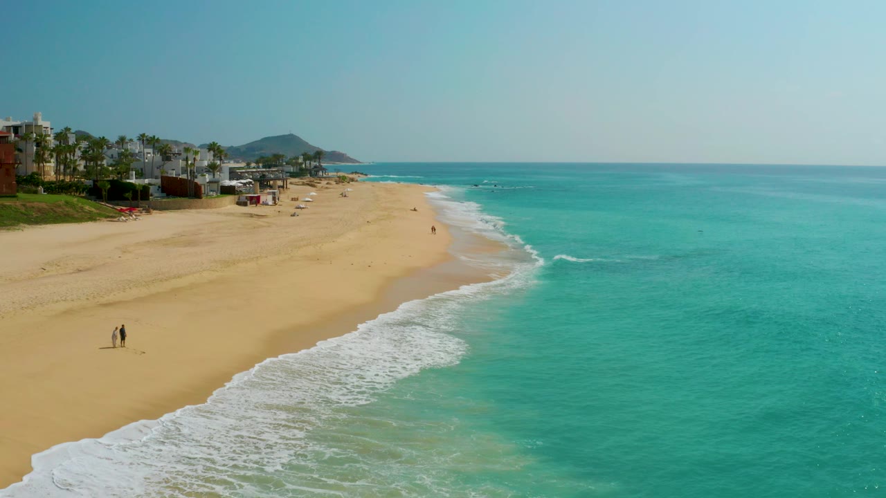 Aerial view over Dreams Beach with a broader sweep of turquoise ocean, foamy surf wrapping the shoreline, resort buildings at the far left, distant hills beyond the coast, and tiny people on the sand.