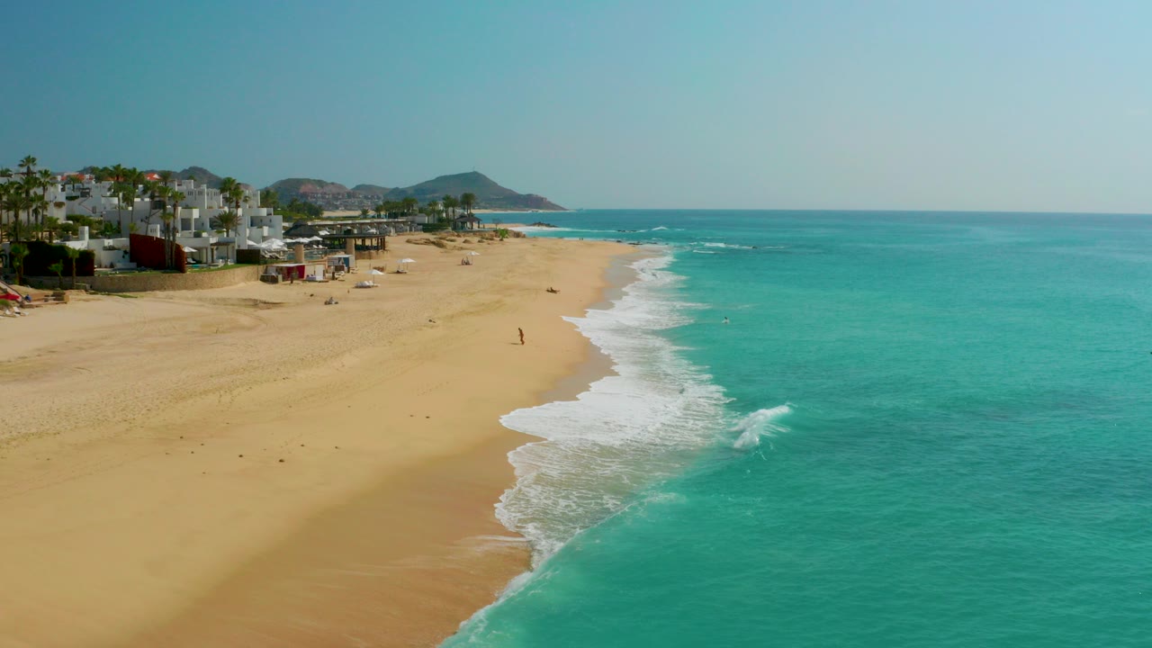 Aerial frame over Dreams Beach in Baja California Sur, Mexico, showing broad sand at left, turquoise ocean at right, white surf curling along the shoreline, and no clearly identifiable people.