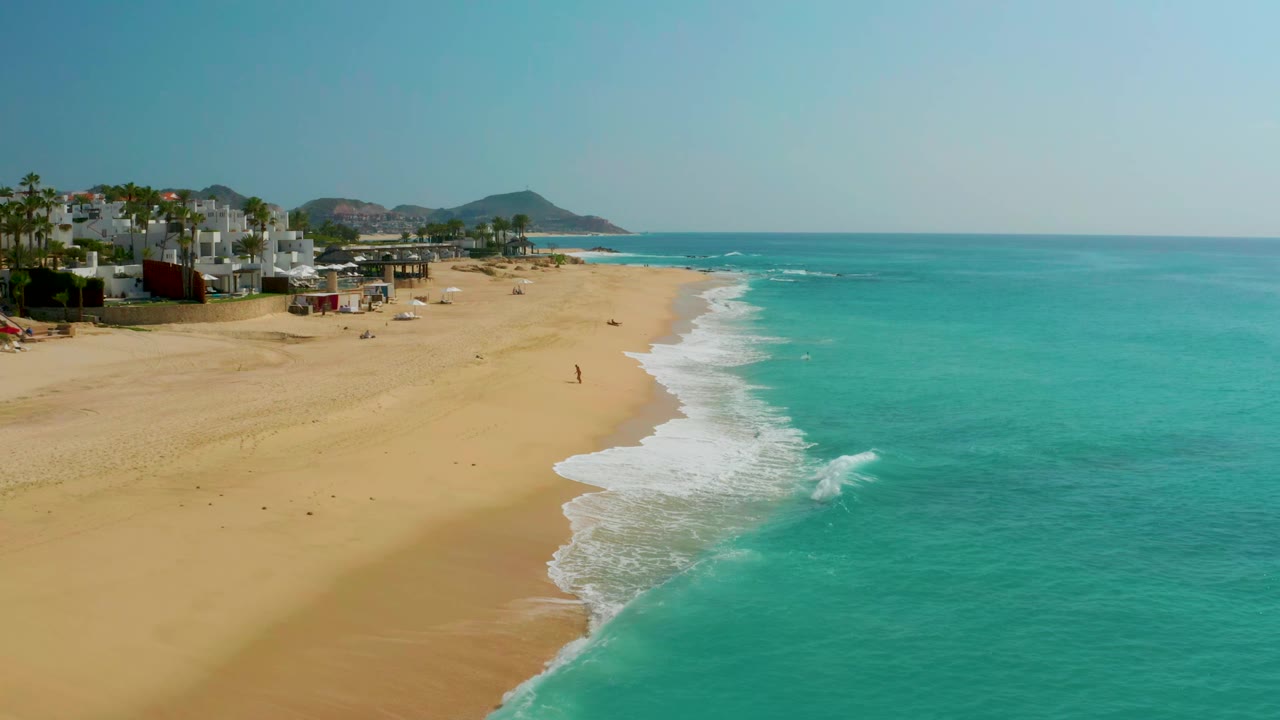 Aerial view of Dreams Beach in Baja California Sur, Mexico, with the surf line slightly farther down the beach, low buildings and palms at left, turquoise water at right, and no clearly identifiable people.