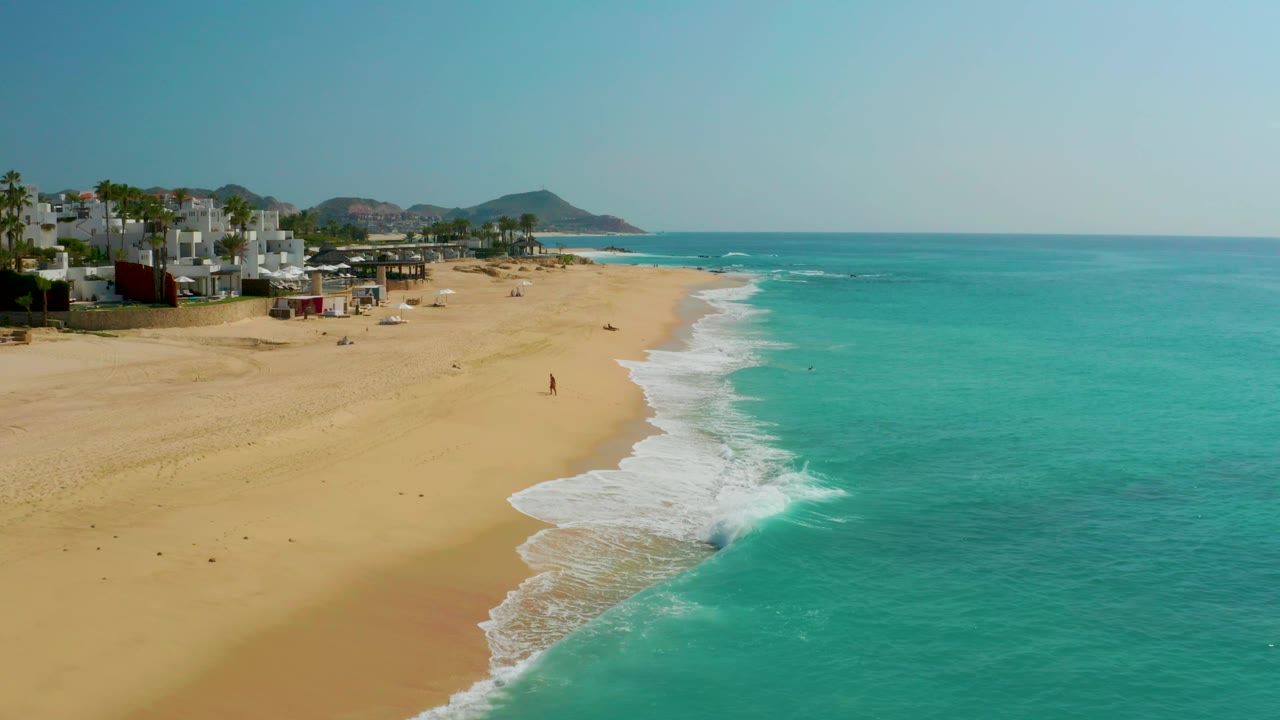 Aerial frame over Dreams Beach in Baja California Sur, Mexico, showing turquoise water, a foamy shore break, resort-edge buildings to the left, distant hills, and tiny beachgoers on the sand.