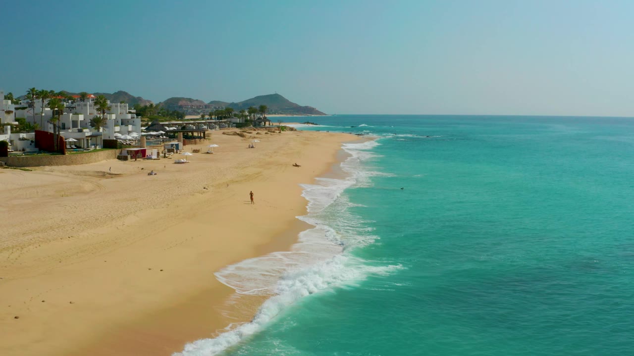 Aerial coast view with buildings, sand, surf, and open water.