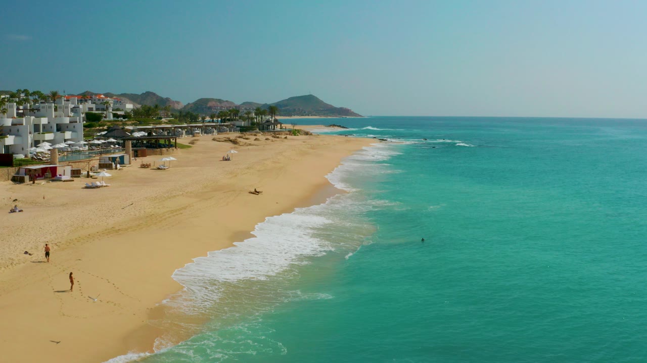 Opening aerial frame over Dreams Beach in Baja California Sur, Mexico, with turquoise water on the right, pale sand on the left, beachfront buildings, distant hills, and tiny beachgoers too small to identify.