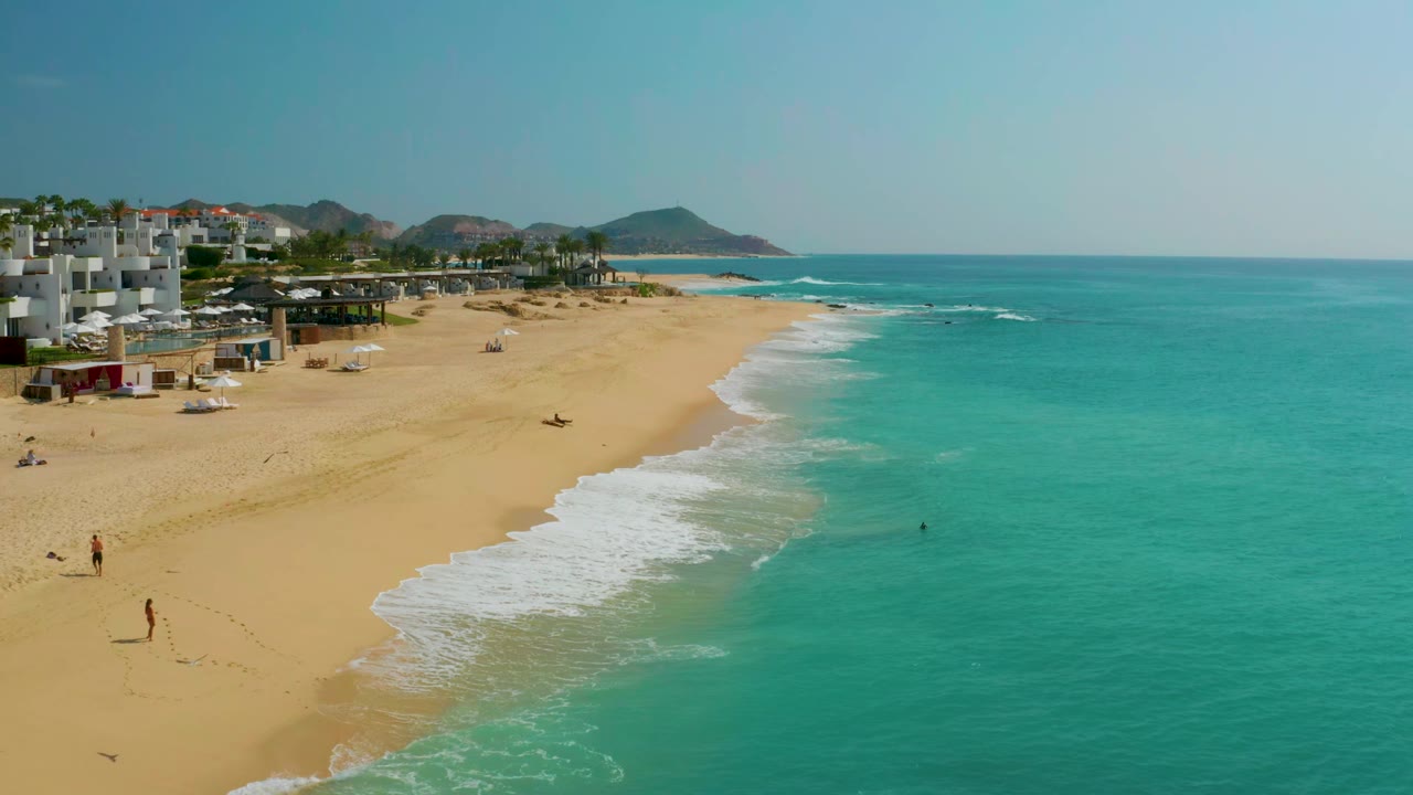 Aerial view one second in, showing Dreams Beach with a diagonal line of white surf, open sand, turquoise water, beachfront buildings, hills, and small distant figures on the beach.