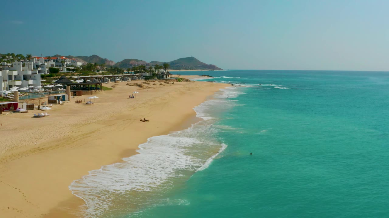 Aerial frame of Dreams Beach with the camera view shifted slightly along the coast, placing more turquoise water in the foreground and the beachfront buildings and low hills along the left background.