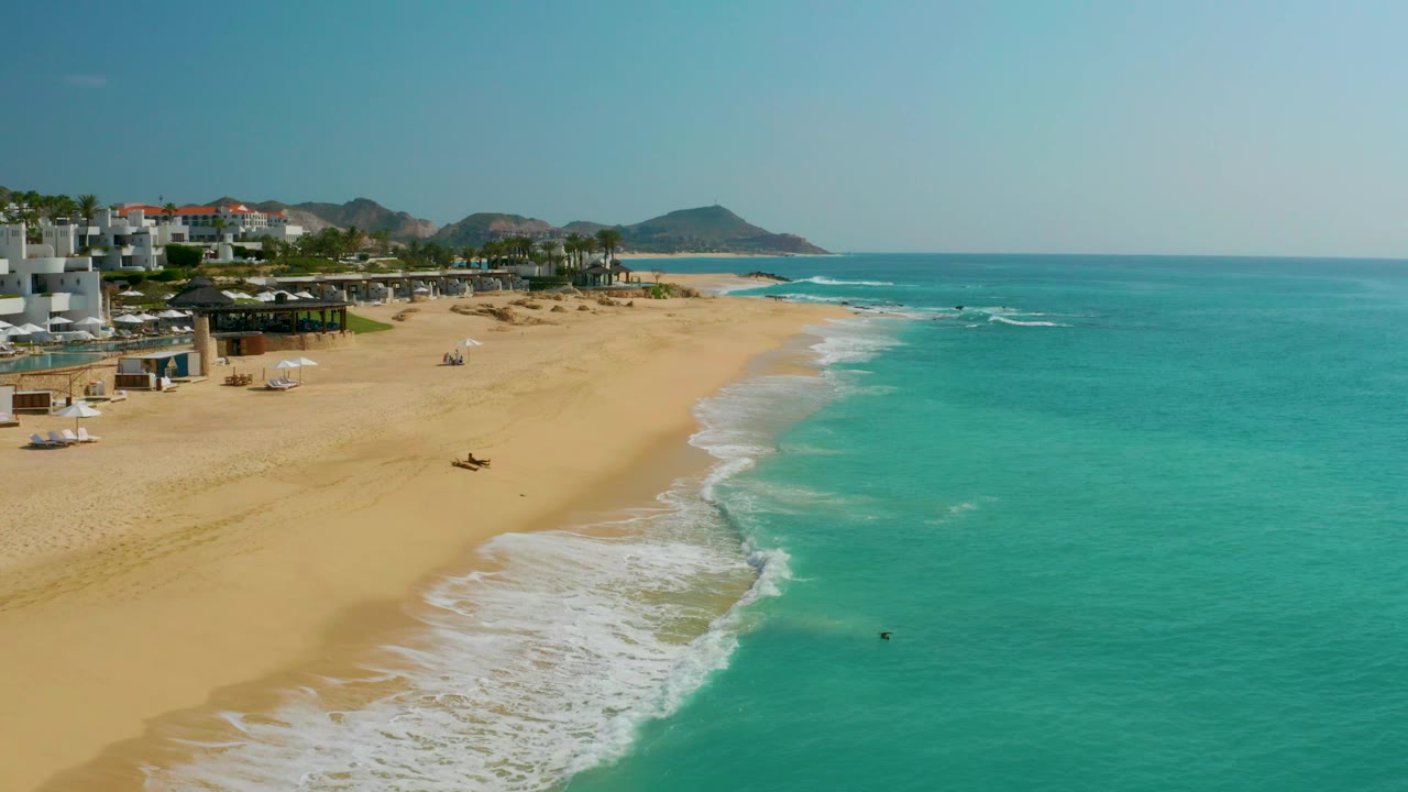 High view of beach, surf, and resort-like buildings under a clear sky.