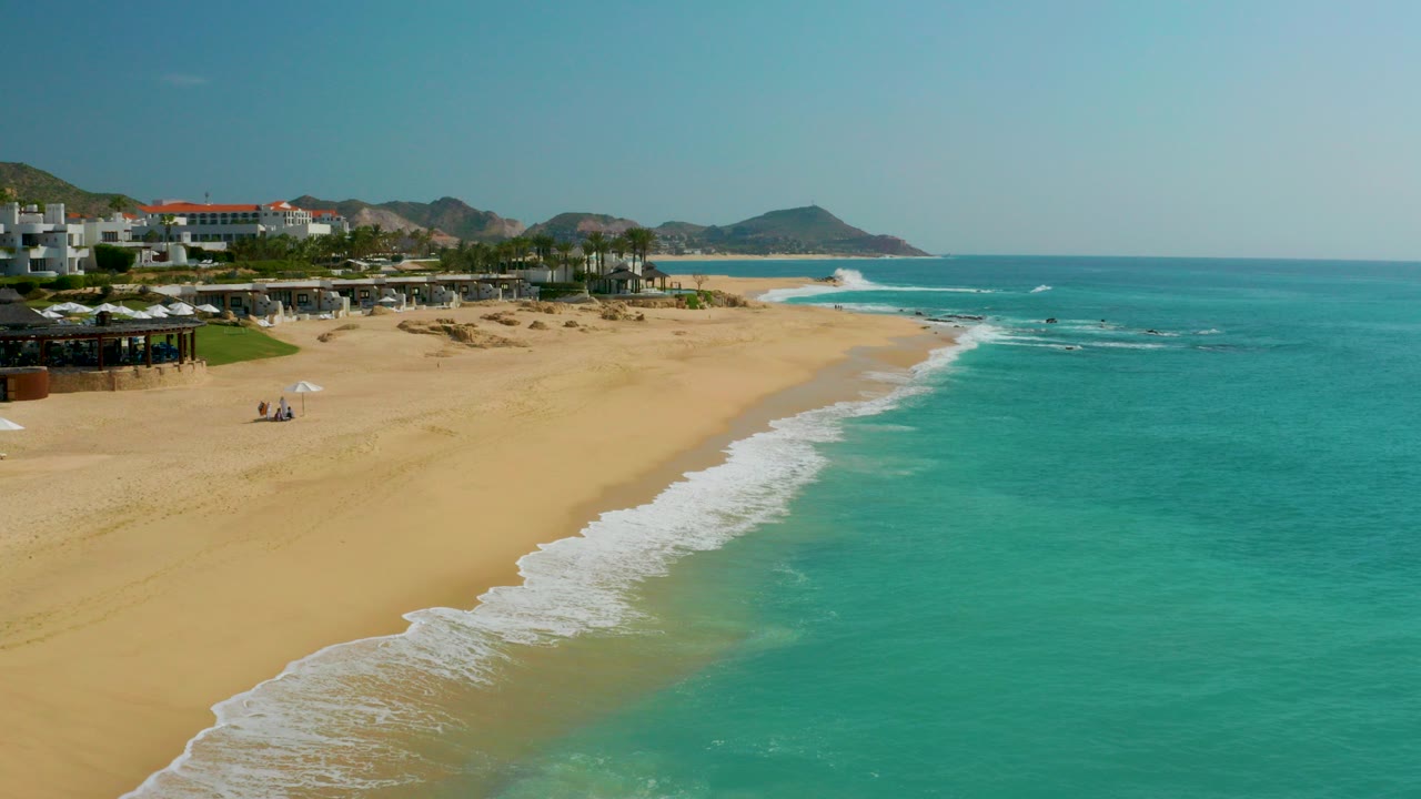 Wide aerial view of Dreams Beach in Baja California Sur, Mexico, with turquoise water on the right, white surf crossing golden sand, beachfront buildings to the left, distant hills, and tiny beachgoers on the sand.