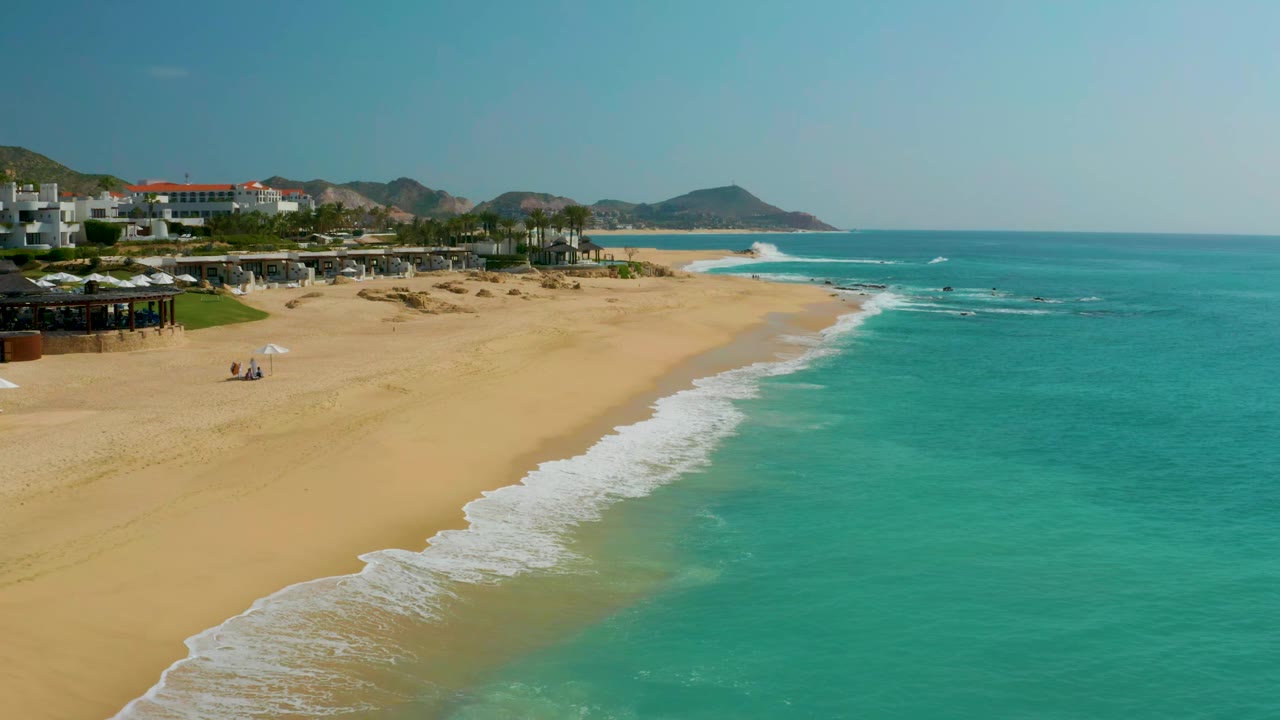 Aerial shoreline view with white waves rolling onto a broad sandy beach.