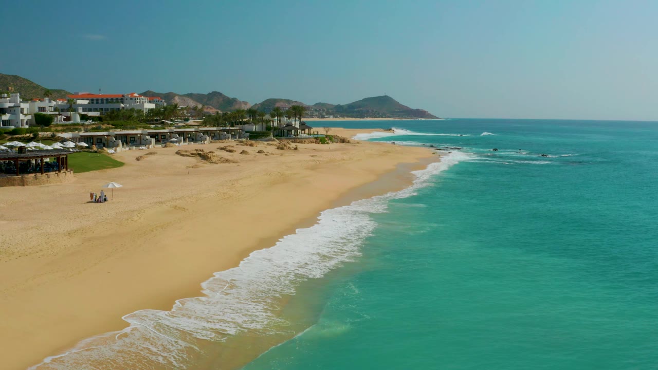 A wide aerial view of Dreams Beach shows the surf line slightly farther along the sand, with turquoise sea filling the right side, beachfront structures at left, distant hills beyond, and tiny beachgoers near the upper beach.