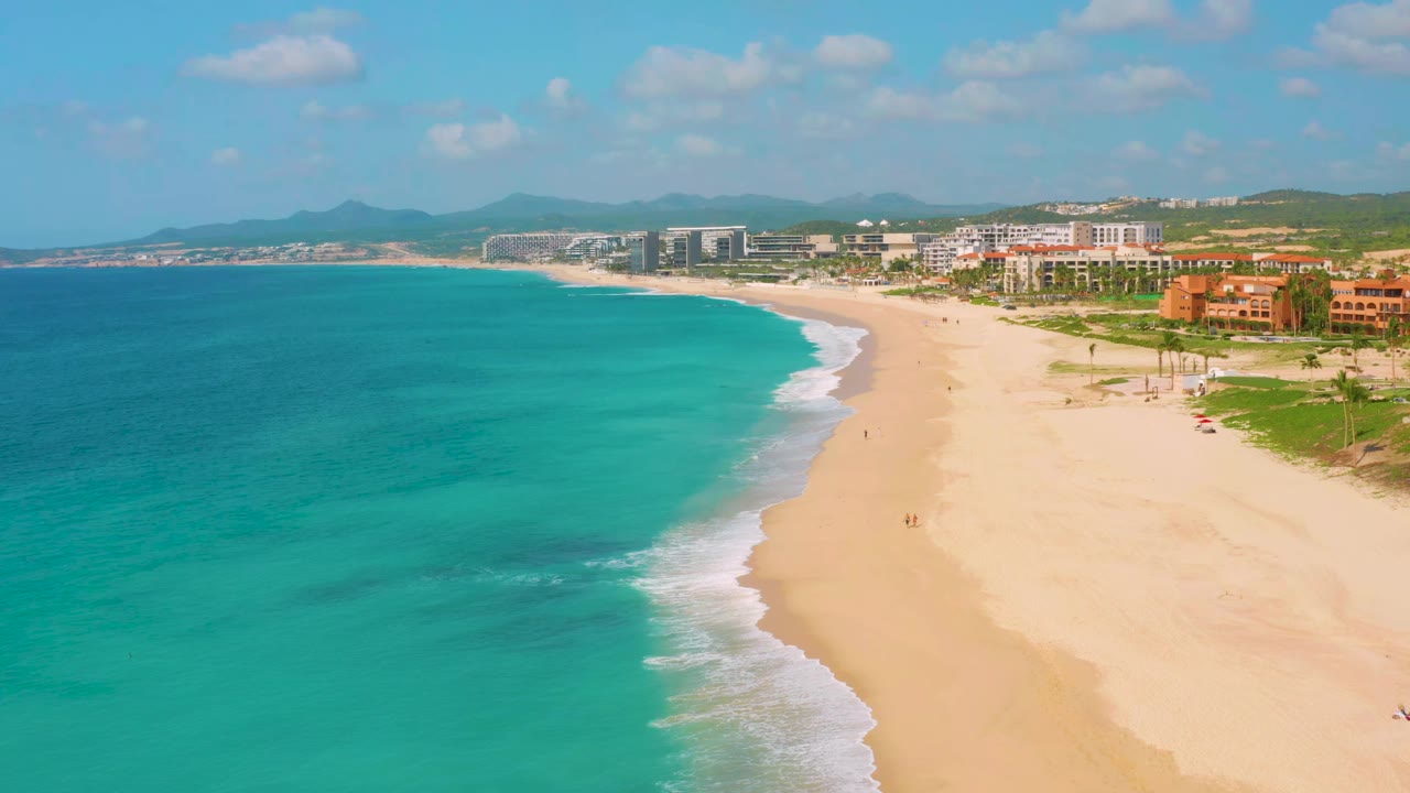 Wide beach curve with blue water, surf, buildings, hills, and scattered clouds.