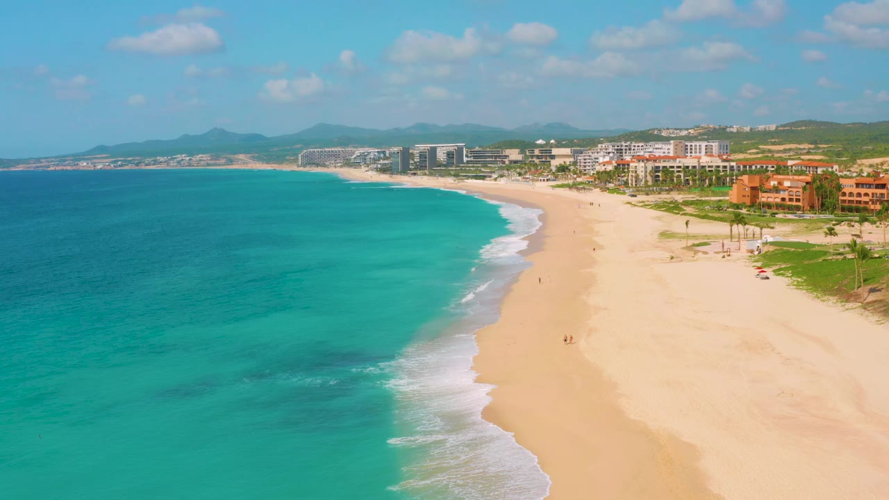 High aerial frame of Dreams Beach with a broad turquoise sea, pale beach, low foamy wave line, resort buildings along the upper right, hills on the horizon, and small distant figures on the sand.