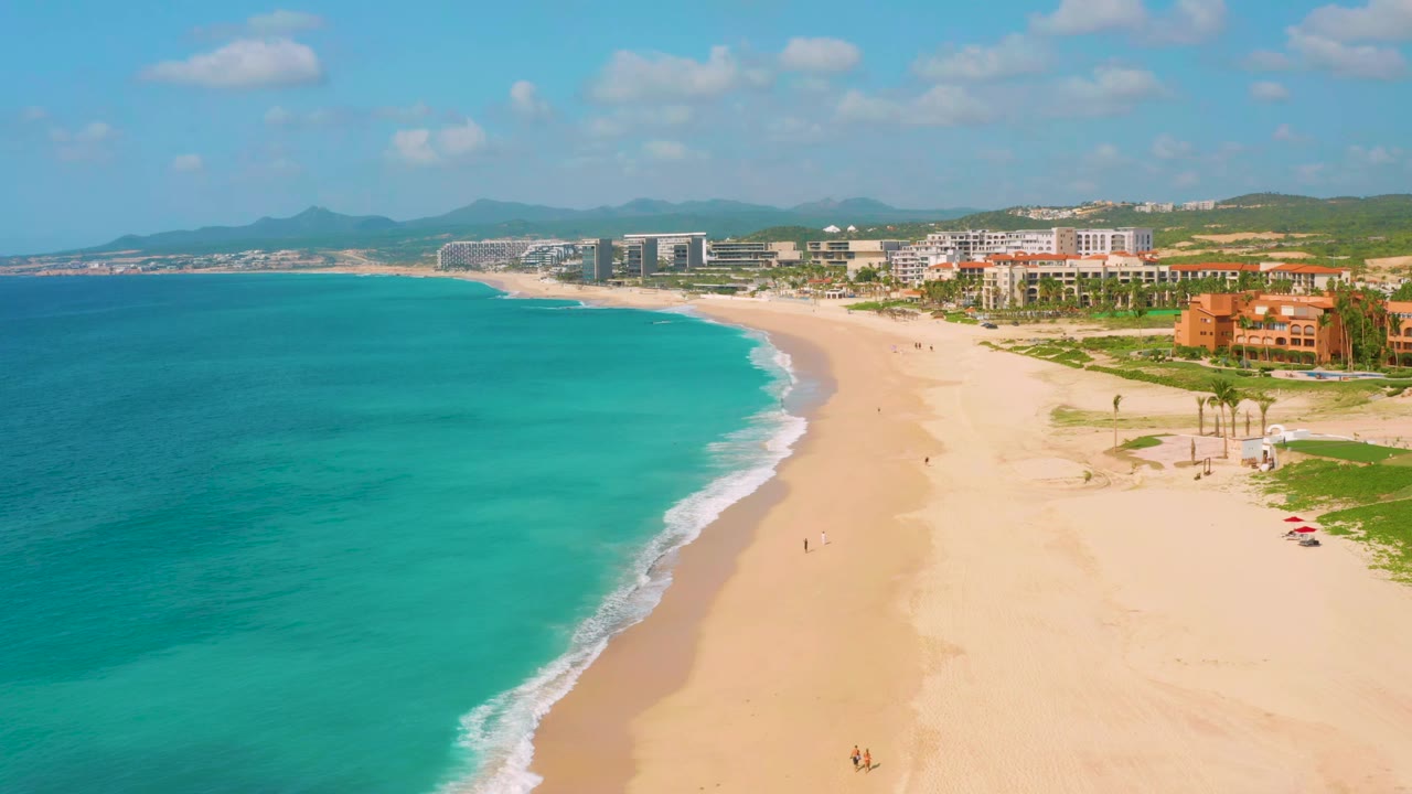 Aerial frame over Dreams Beach showing the surf line running diagonally through pale sand, with turquoise water, resort blocks, green inland areas, and hazy mountains beyond.