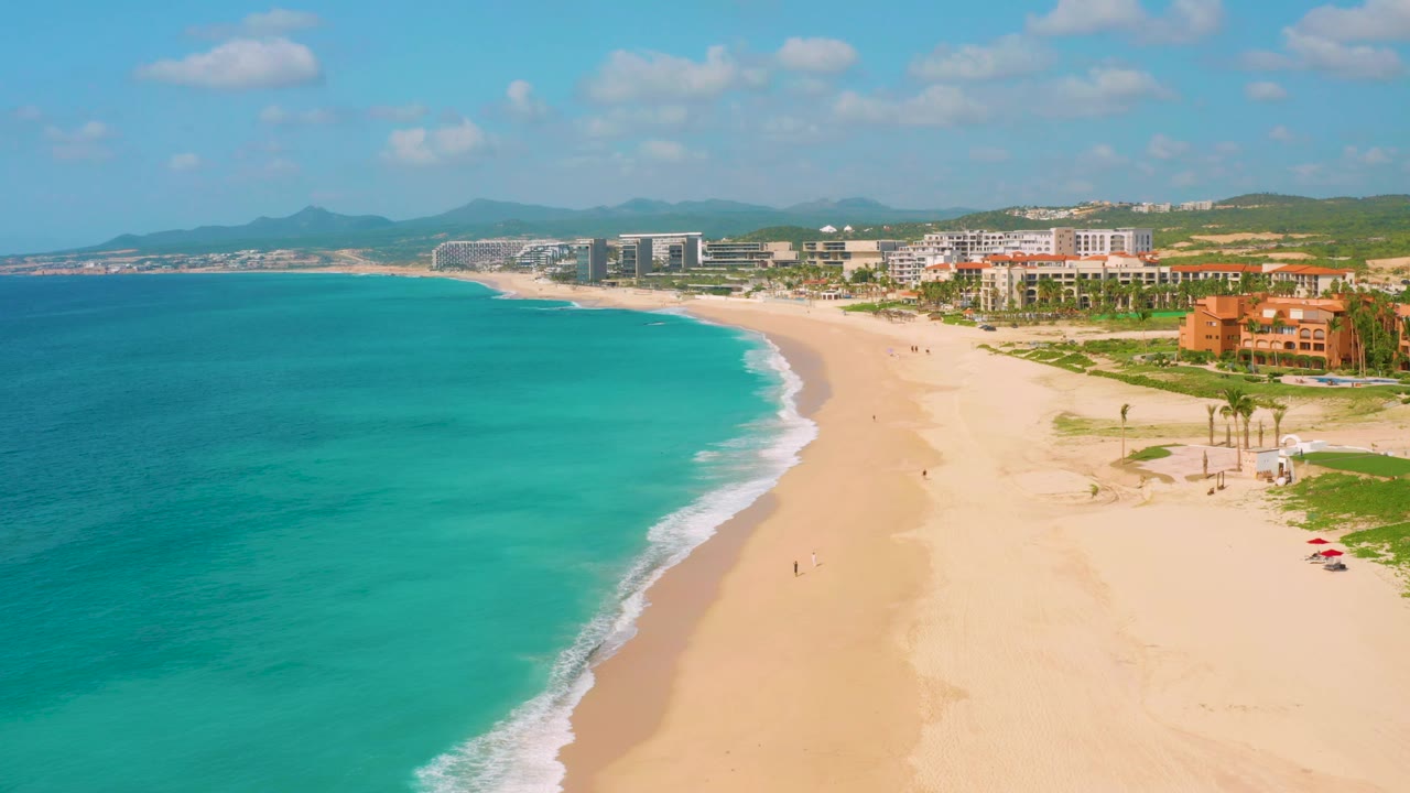 Aerial frame at Dreams Beach with more of the sandy curve visible, turquoise water occupying the left half, resort buildings along the upper right, and small distant marks on the beach that may be people.