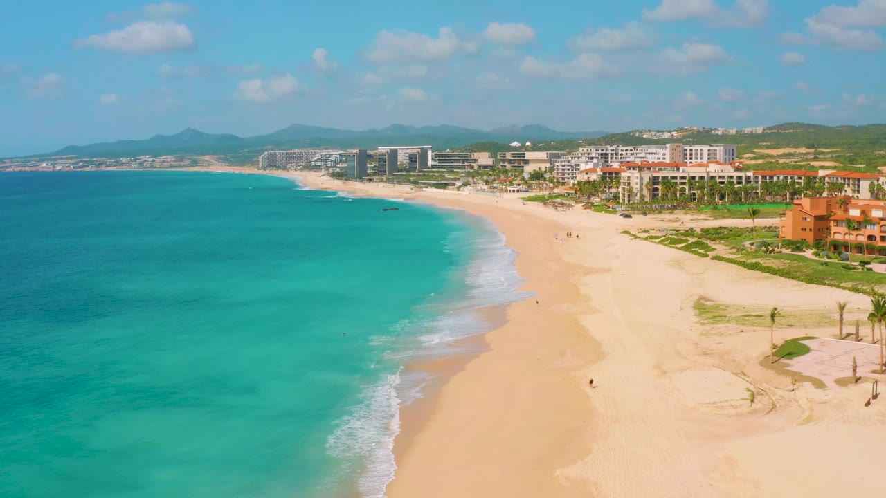 Dreams Beach aerial view showing a longer curve of pale sand, shallow blue-green water, small breaking waves, and a line of beachfront buildings in the distance.