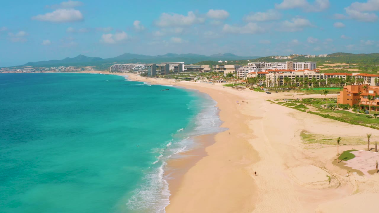 A wide aerial over Dreams Beach with the surf line running diagonally across the frame, turquoise sea to the left, open sand to the right, and coastal buildings along the background.