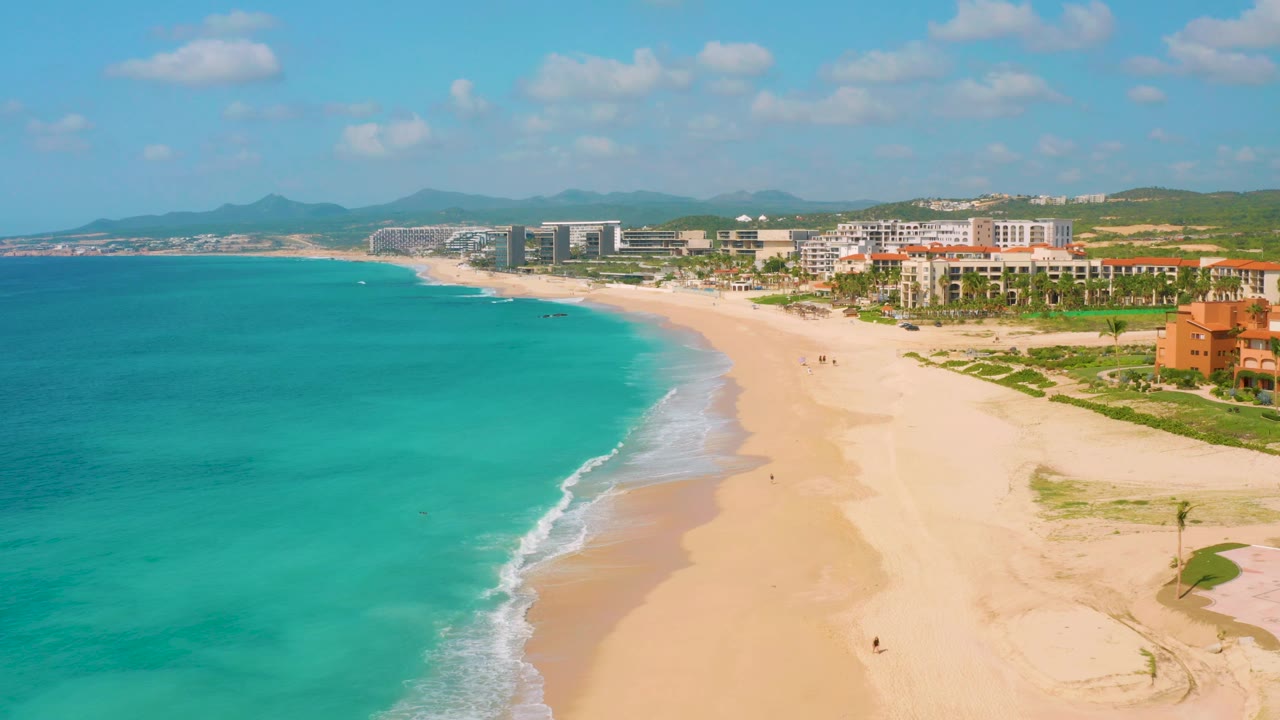 Drone view of a curving beach with turquoise water and distant development.