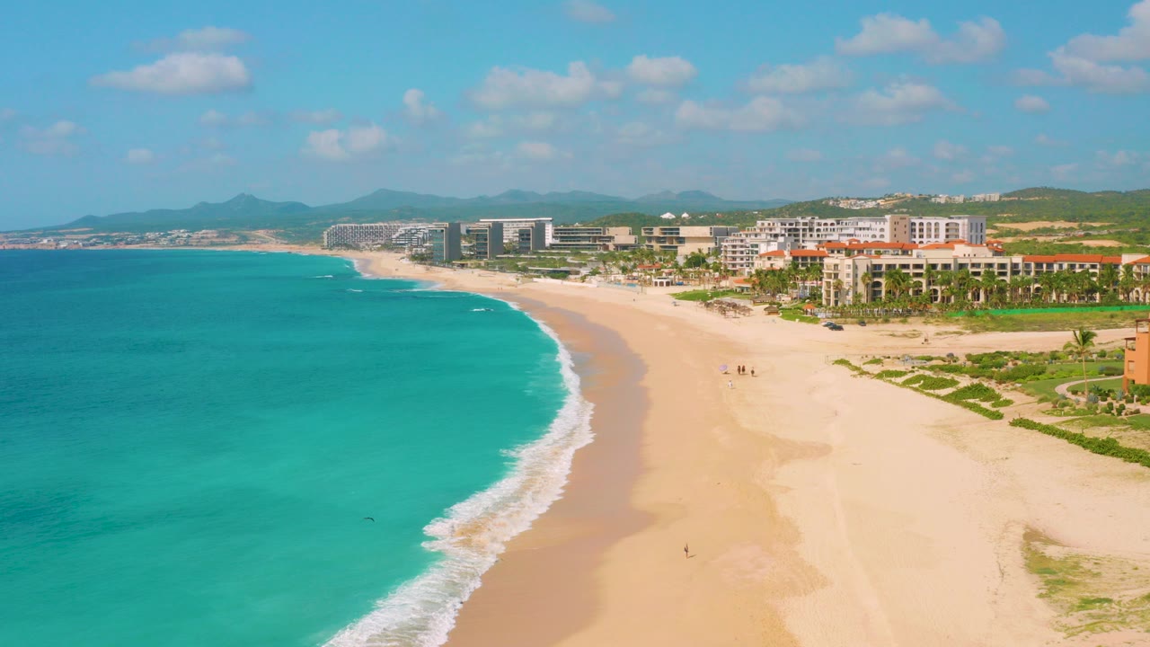 Aerial view over Dreams Beach in Baja California Sur, Mexico, with turquoise water at left, a curving surf line, broad sand, beachfront buildings, distant hills, and tiny distant beachgoers on the shore.