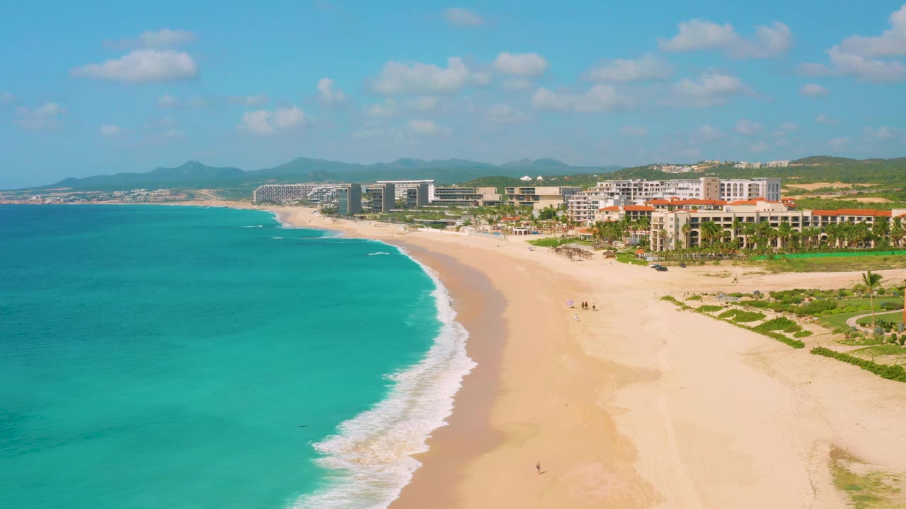 At 0:02, the aerial view shows a broad sweep of Dreams Beach, with teal water on the left, pale sand on the right, a thin foam edge, and beachfront buildings backed by green hills.