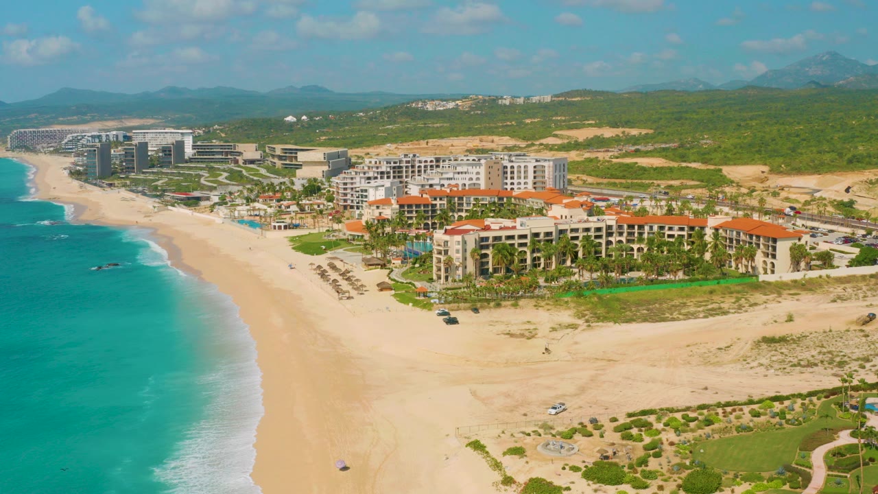 Opening frame over Dreams Beach in Baja California Sur, Mexico, with turquoise water at left, pale sand in front, resort buildings beyond the beach, green hills behind, and no visible people.