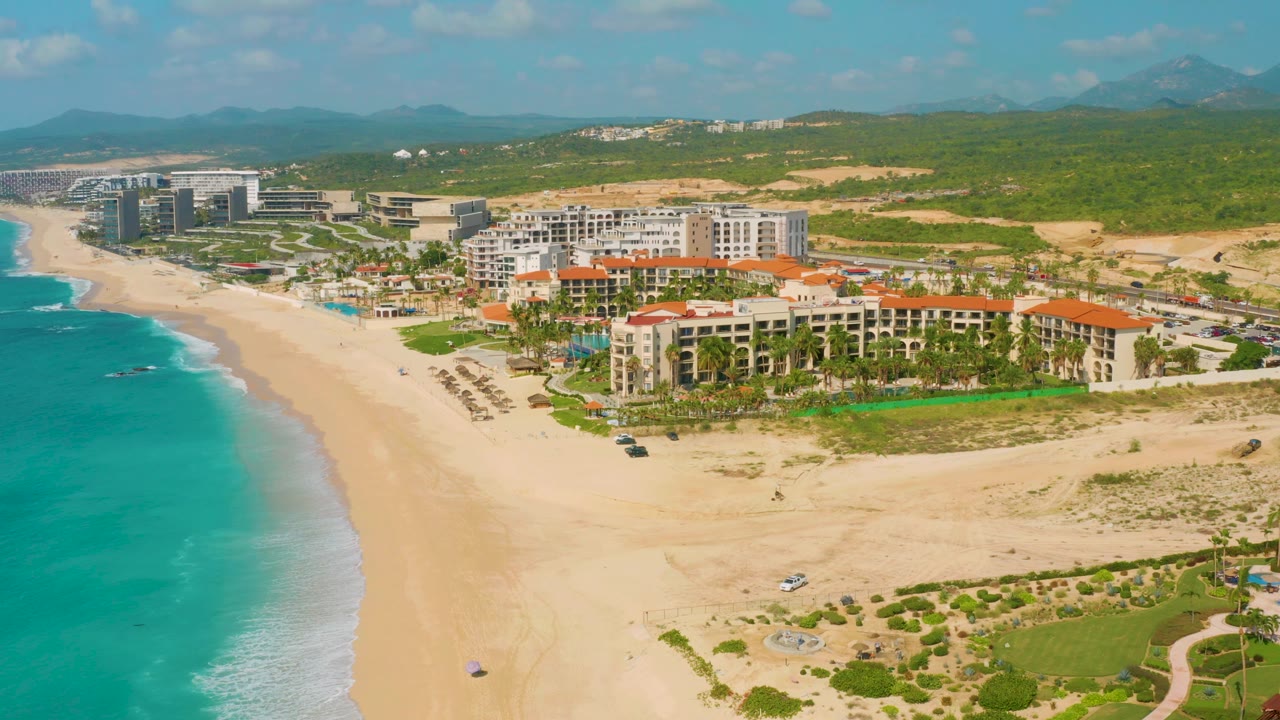 Fourth frame places the Dreams Beach resort slightly right of center, with foamy waves along the left shoreline, open sand in front, green hillside behind, and no visible people.