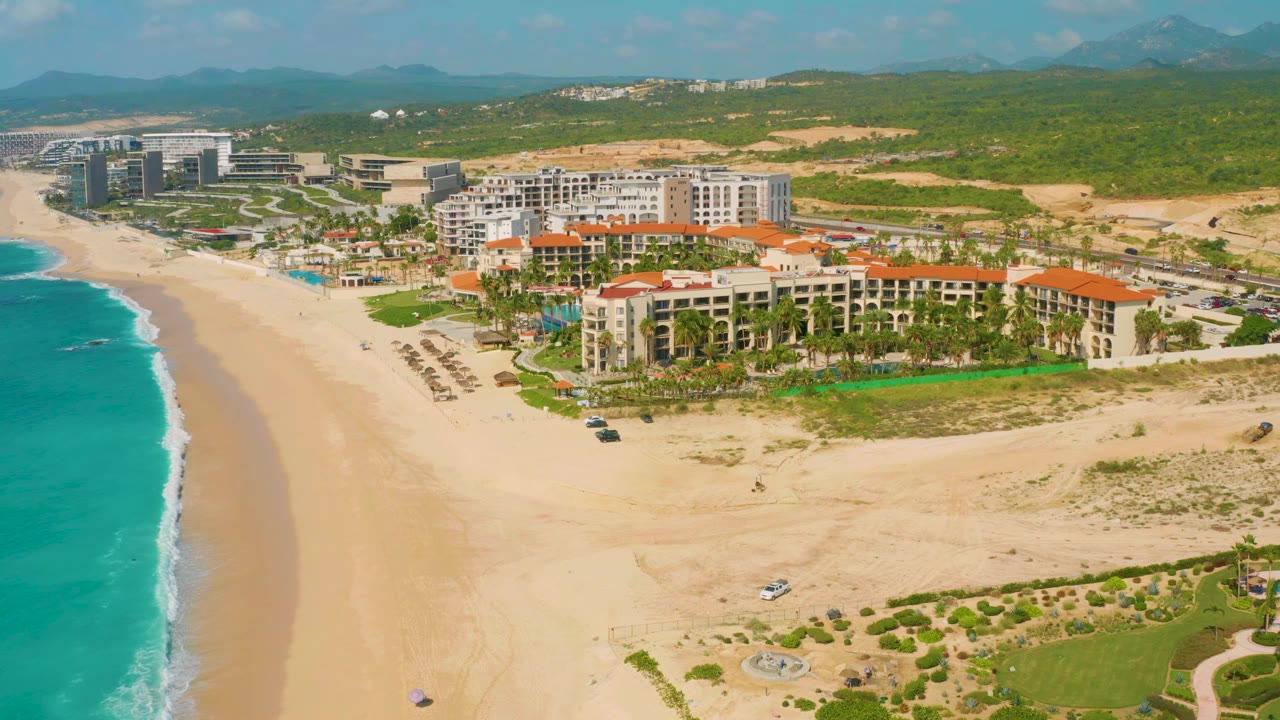 Elevated drone view of the Baja California Sur beach resort, with the surf line tight along the left edge, pale sand below, clustered resort buildings in the center, and no visible people.