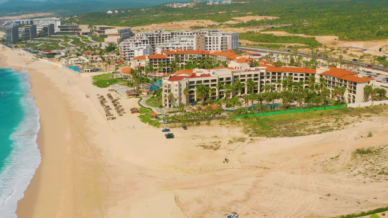 Aerial frame in Baja California Sur, Mexico, with the orange-roofed resort centered above a broad sandy beach, turquoise water on the left, and no visible people.