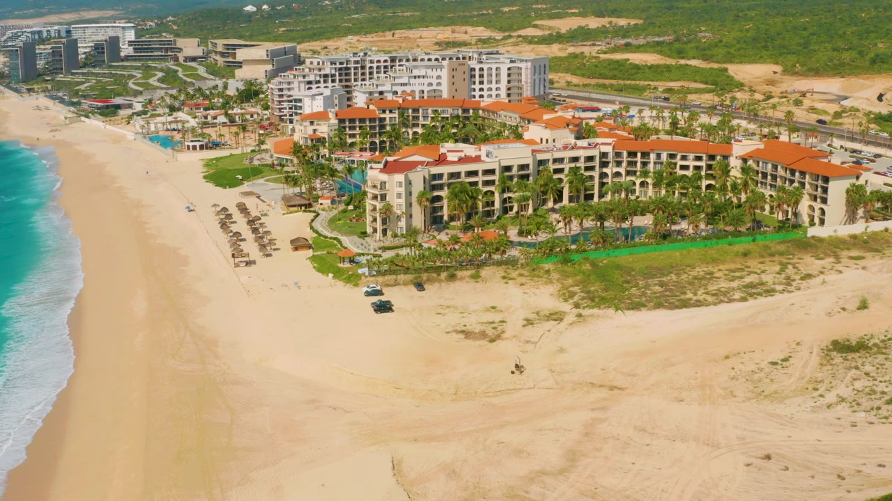 Aerial view in Baja California Sur, Mexico, showing the resort slightly closer, with pool water, palm-lined grounds, rows of beach shade structures, and no visible people.