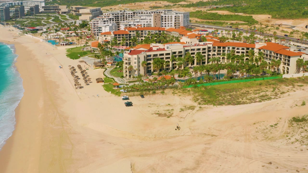 A closer aerial frame in Baja California Sur, Mexico, with the resort filling more of the right side, surf and sand on the left, parked vehicles near the beach, and no visible people.