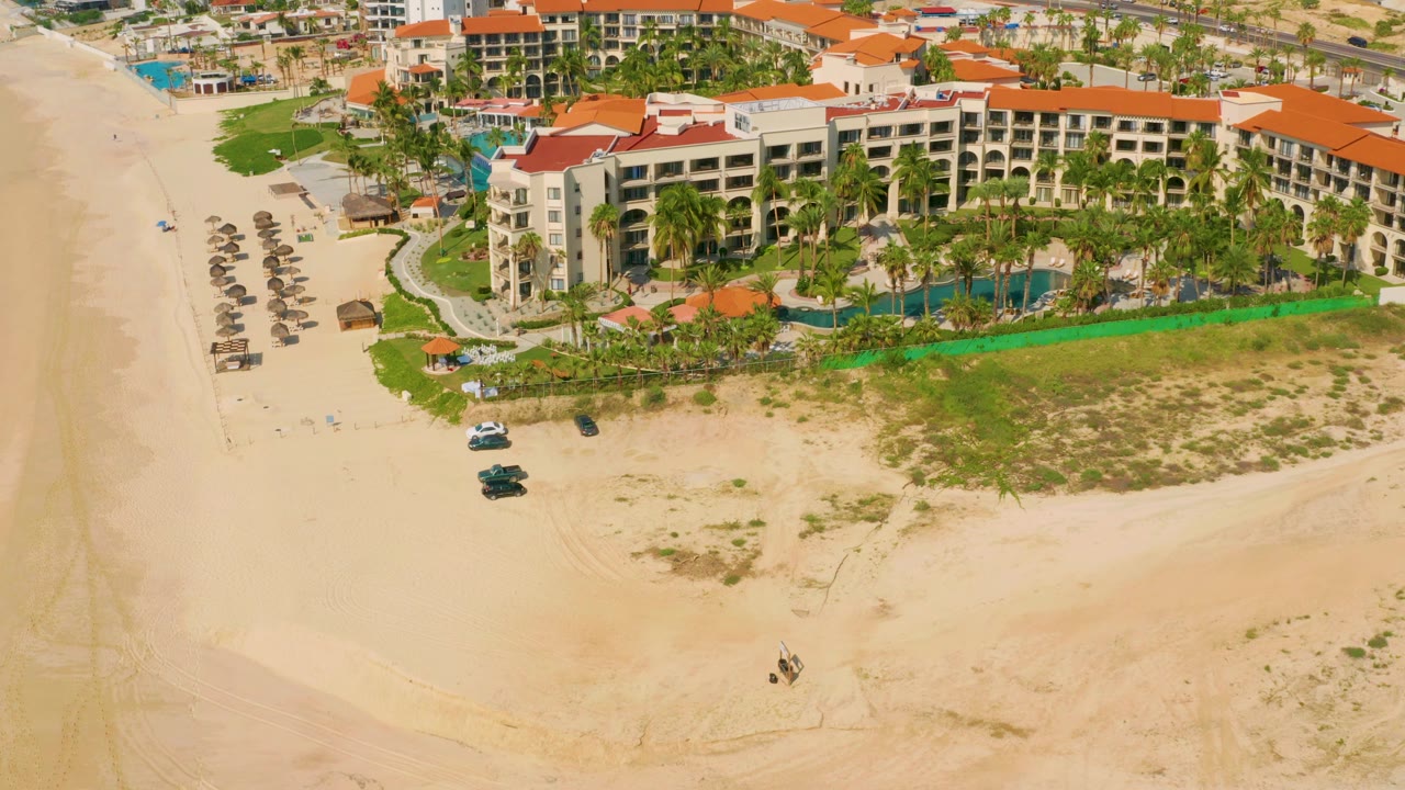Aerial view at 0:01 over Dreams Beach in Baja California Sur, Mexico, with the resort building centered, rows of beach shade structures at left, sand below, and no visible people.