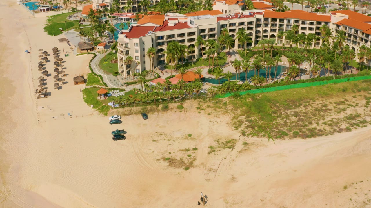 Mid-clip aerial frame over Dreams Beach, Baja California Sur, Mexico, with the hotel facade closer in view, blue pools behind palms, sandy beach below, and no visible people.