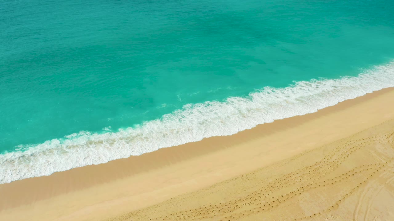 Overhead shoreline at Dreams Beach where blue-green water presses a textured white foam edge onto smooth golden sand, with no visible people.