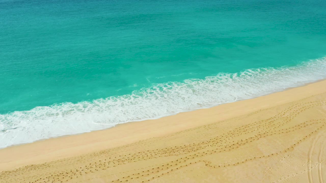 Aerial view of turquoise surf reaching a pale sandy beach at Dreams Beach in Baja California Sur, with dotted footprints across the sand and no visible people.